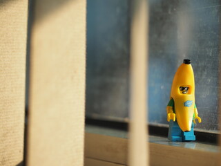 Fototapeta premium Indonesia - September 12, 2025 : Mini toy of action figure at table with blurred background. Miniature photography concept design. Minifigure of banana guy character at office window with sunset light