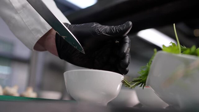 Close up of chef hands in black gloves slicing mushrooms with knife into white bowl. Action moves from slicing to finishing. Professional kitchen setting with green herbs nearby