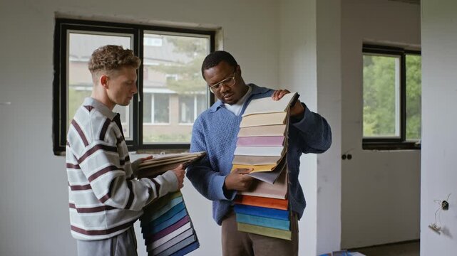 African American young adult man and Caucasian young adult man choosing fabric swatches in unfinished room. Footage suitable for interior design and home renovation promotions