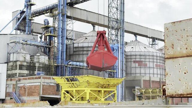 discharging wheat from ship by crane grab to hopper at industrial port with tank and conveyor belt.