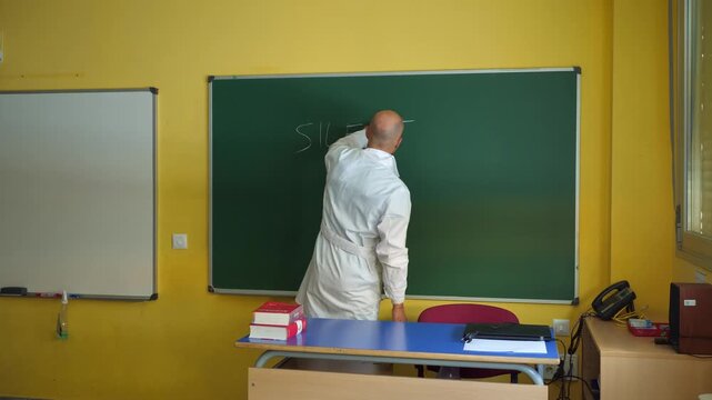 Medium wide shot of a male teacher in a white lab coat writing 'SILENCE!' with chalk on a green chalkboard. He turns around with a serious expression, grabs a yellow megaphone from his desk and shouts