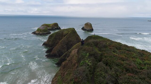 A cinematic drone shot fly's low over hikers on a cliffs edge on sea spires known as "Bird Rocks" rising from the Pacific beside John Yeon State Natural Site at Chapman Point, on Oregon&rsquo;s coast.