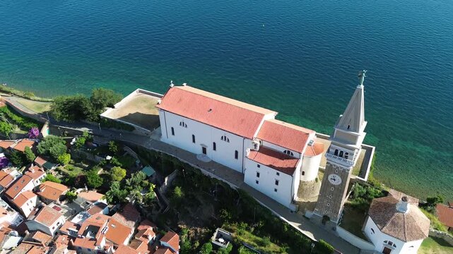 Drone aerial view of Piran old town with St. George Church, red tiled rooftops and Adriatic Sea coastline on a sunny summer day in Slovenia.