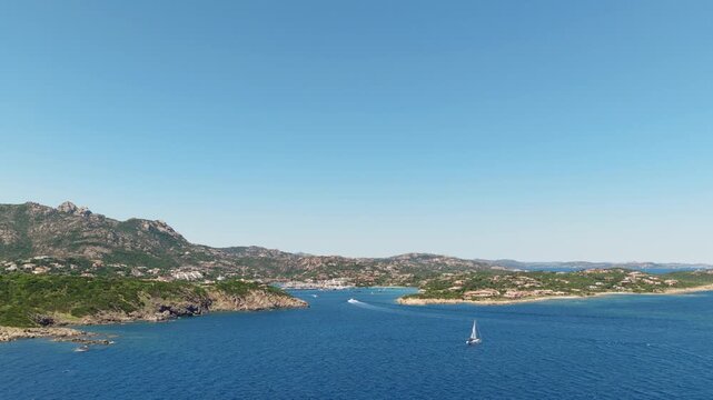 Sailboat moves across the deep blue bay of Porto Cervo in Sardinia, framed by rocky hills and coastal villas under clear skies.