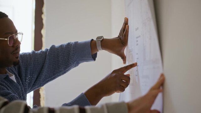 African American man and Caucasian man pointing at blueprint on wall, discussing home renovation plans. Clip is suitable for real estate marketing and construction planning commercials
