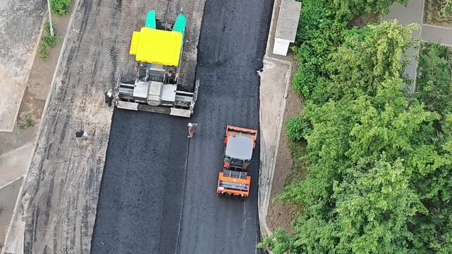 Construction workers operate heavy machinery to lay asphalt on a road, showcasing the continuous process of paving with a yellow and orange roller in a green urban environment