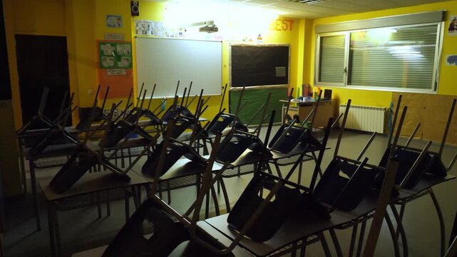 Wide angle shot of an empty and quiet primary school classroom during low light or night. Rows of student desks with chairs stacked upside down fill the room. Concept for school break or end of class