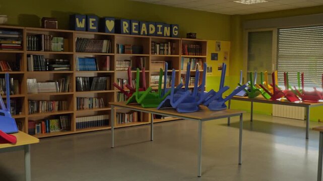 Panning shot of an empty primary school library with bright colorful chairs stacked upside down on tables at the end of the day or summer break.