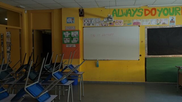 Slow panning shot of an empty and dimly lit classroom showing desks with chairs stacked upside down. A whiteboard displays the motivational text 'DO IT ANYWAY'