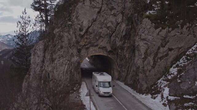 Frontal drone shot of a white camper van exiting a narrow rock tunnel on a mountain road in winter, Tara River Canyon, Montenegro. Concept for road trip journey.