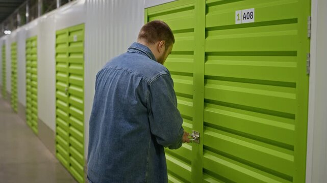 Caucasian young adult man unlocking padlock on green door of sel storage unit. Video is perfect for commercial ads promoting moving services, decluttering, or personal storage solutions