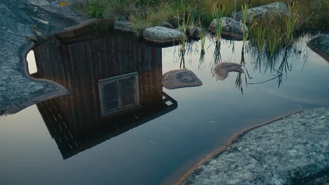 Abstract reflection of a weathered wooden boathouse mirrored in a calm tidal pool surrounded by smooth coastal granite and shoreline vegetation. Peaceful Scandinavian waterscape with natural textures.