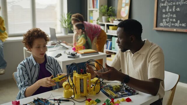 African American male teacher explaining robotics to Caucasian child holding controller in STEM classroom. Footage suitable for promoting modern school technology programs and science education