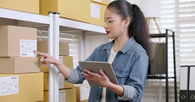Asian woman entrepreneur points toward parcel label while checking order data on tablet coordinating e-commerce inventory management and online store order fulfillment inside warehouse space