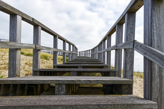 Boardwalk steps with railings climbing across a sandy dune under a cloudy sky, forming a path to the top
