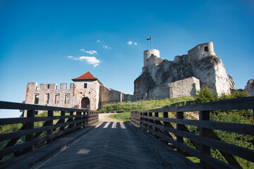 A wooden bridge leading to ancient medieval stone castle ruins situated on a rocky hill under a clear bright blue summer sky with a waving flag. © Bogdan