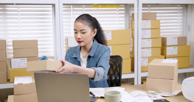 Asian woman entrepreneur holding parcel box while scanning shipping label, checking order details, reviewing inventory status, managing online shopping parcel box workflow inside organized home
