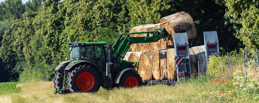 Green tractor loads round hay bale onto farm trailer stacked with straw beside meadow. Summer harvesting scene shows rural machinery, agriculture, transport, fieldwork, and sunny countryside