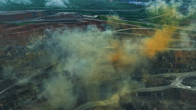 Aerial tracking shot of open pit mine with dust and orange smoke after detonation plumes over terraced excavation roads, mining industry concept.