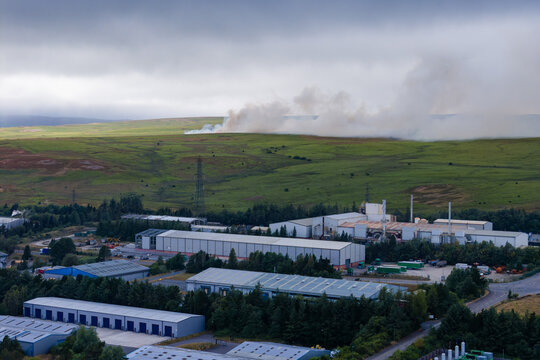 Smoke plume from wildfire rising over dry moorland above industrial estate, high angle view of grassy hill slopes under overcast sky near Ebbw Vale