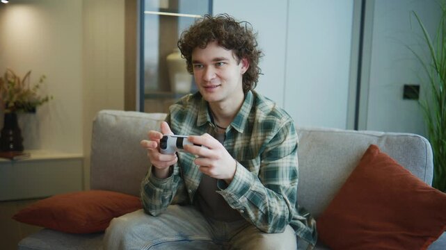 A young man sits on a sofa in a modern room, holding a game controller and focused on the screen. Daylight fills the space through the window.
