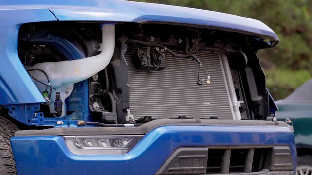 Modern blue pickup truck with significant front-end damage sitting in a car lot, its missing grille exposing the radiator and engine components while awaiting collision repair or salvage