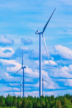 Three wind turbines spinning against a bright blue sky with fluffy white clouds above green trees in Finland