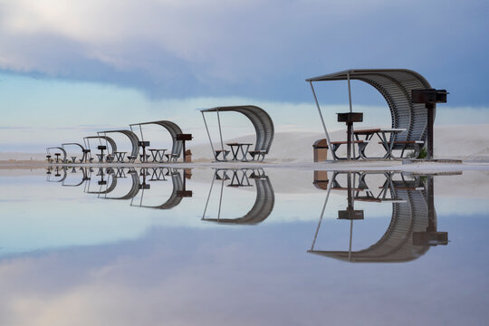 Rows of curved sun shelters, cabanas with seats for visitors to the White Sands national park reflected in the shallow surface water.