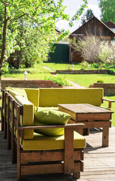 Wooden pallet sofa with green cushions on sunny garden terrace. Relaxing outdoor living space with green grass and trees