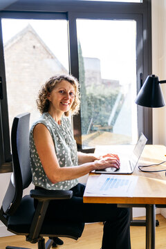 Smiling woman sitting at desk, using laptop for business work in a contemporary coworking office space