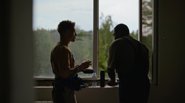 African American and Caucasian young adult builders standing by window, eating lunch and drinking coffee during break. Footage is perfect for home renovation and construction services marketing