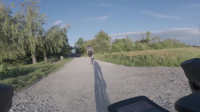 POV first person view of a gravel cycling adventure in Germany following another rider on a sunny country dirt road fork surrounded by green trees and fields during a long distance bike trip
