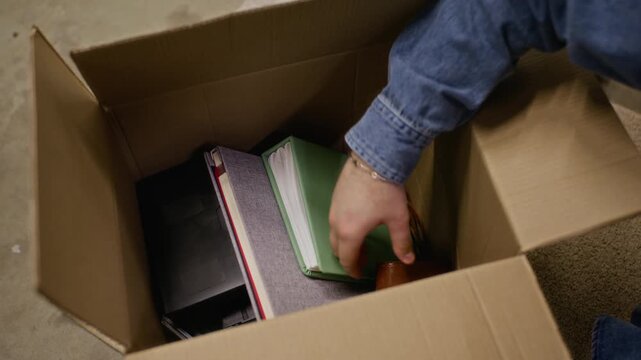 Unrecognizable young adult unpacking ukulele, vintage camera, and VHS tapes from cardboard box in storage unit. Video is perfect for moving, decluttering, or nostalgia themed marketing campaigns