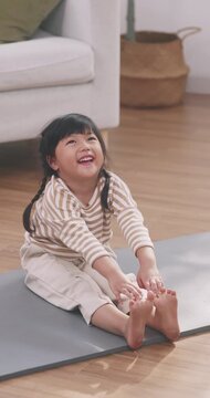 Cute Asian little girl stretching her body and touching her feet on a yoga mat at home.