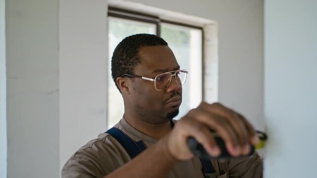 African American young adult handyman in overalls measuring wall with tape measure in unfinished room. Video is perfect for home renovation services, DIY tutorials, and construction marketing