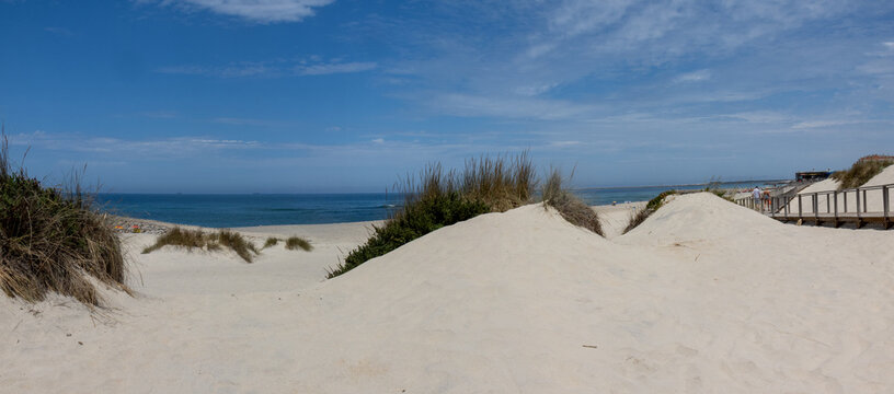 Wooden boardwalk leading through the white sand dunes to Costa Nova Beach near Aveiro, Portugal. A peaceful coastal scene with protected dunes, clear blue sky, and direct access to the Atlantic Ocean.