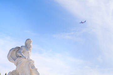 Fototapeta premium Ancient marble deity statue side profile against a pale blue sky, with an airplane passing overhead.