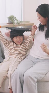 Cute little Asian girl wearing graduation cap holding diploma and celebrating with her mother on the sofa at home.