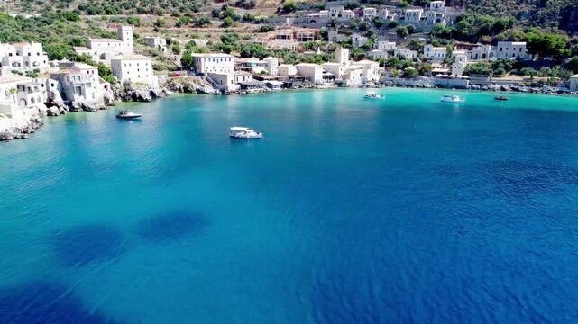 Drone footage of traditional fishing boats floating in the crystal-clear turquoise waters of Limeni Bay, Mani, Greece, with historic stone buildings along the Mediterranean coastline.