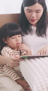 Asian mother sitting on bed reading a storybook with her cute young daughter at home.