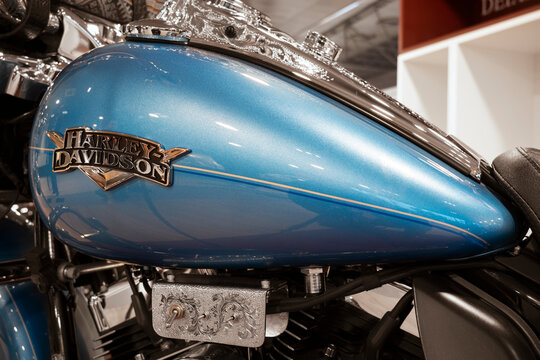 Close-up of a metallic blue Harley-Davidson fuel tank with chrome emblem and custom engraved details at a motorcycle exhibition.