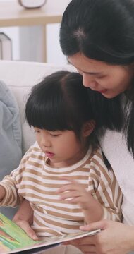 Asian mother sitting on bed reading a storybook with her cute young daughter at home.