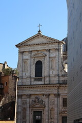 Obraz premium A historic Baroque church facade with a cross on top, stone carvings, and Latin inscription stands between surrounding buildings under a clear blue sky.