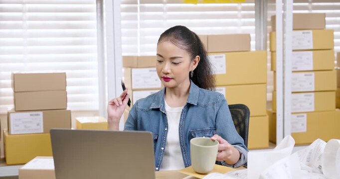 Asian woman small business owner reviews online orders on laptop while holding pen and coffee cup checking documents during e-commerce packaging routine inside home warehouse with stacked parcel boxes