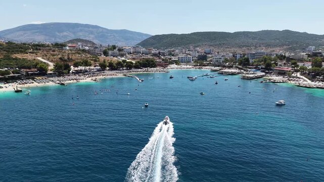 Aerial top-down drone view of a speedboat cruising through the vibrant turquoise waters of the Ionian Sea, leaving a white foamy wake. Beautiful crowded sandy beach of Ksamil coastal resort town with 