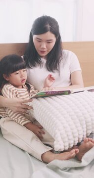 Asian mother sitting on bed reading a storybook with her cute young daughter at home.