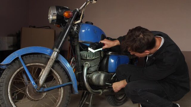 Man focusing on repairing the engine of an old blue motorcycle with a flashlight in a dimly lit garage, performing maintenance or restoration on the classic vehicle