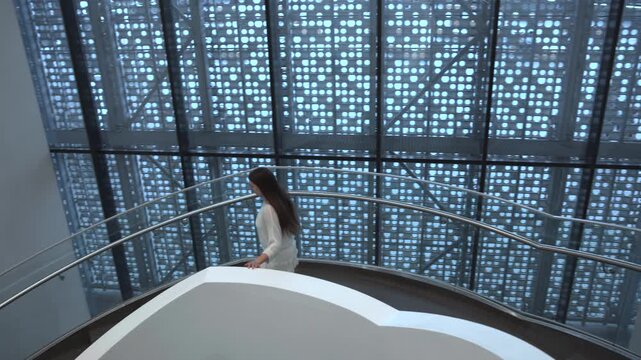 Woman descending curved staircase in modern atrium with perforated metal facade and glass balustrade, overhead oblique viewpoint showing contemplative pose and hand on railing