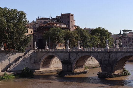 An ancient stone bridge with marble angel statues spans a muddy river with historic buildings and trees visible in the background.