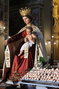 A ornate statue of the Virgin Mary wearing a golden crown holds the infant Jesus surrounded by lit votive candles inside a church.